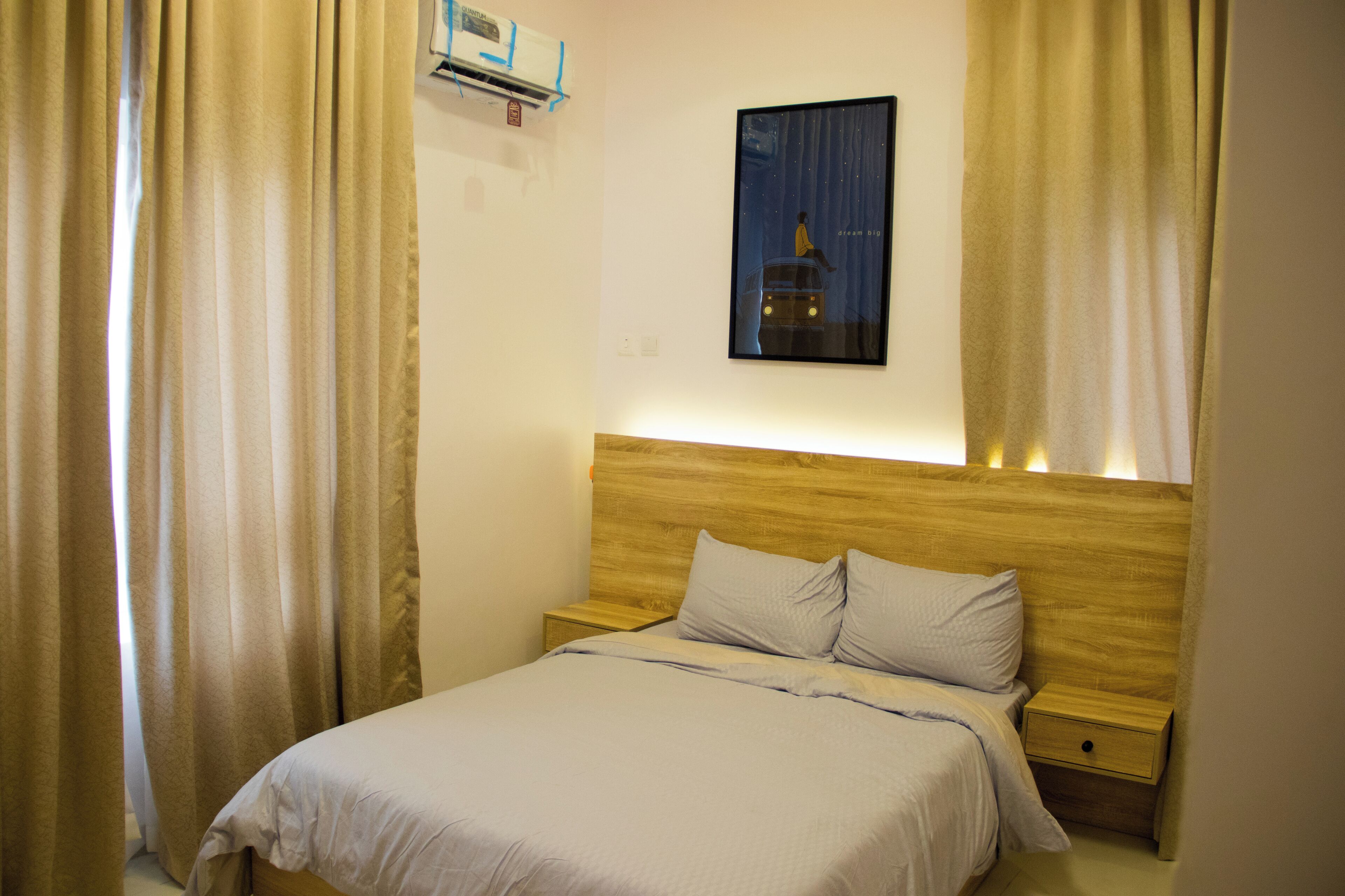 A bedroom with a wooden headboard, a single bed with a white duvet, two bedside tables, and a wall-mounted air conditioning unit.