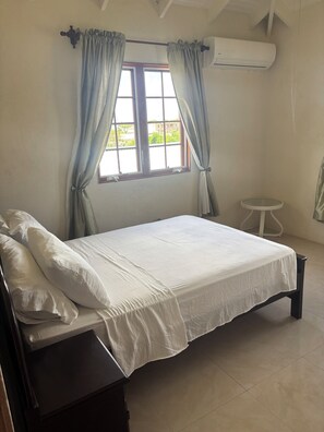 A bedroom with a bed, curtains, a window, an air conditioning unit, and a small table. - Ealing Park South Apartments in peaceful Christ Church with AC (Christ Church)