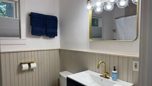 Un baño con inodoro blanco, lavamanos blanco con grifo dorado, espejo con luces y toalla azul en la pared.