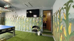 Una habitación con proyector, televisor, una mesa, una silla y un mural de mazorcas de maíz en la pared.