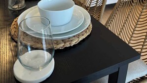 Ein Esstisch mit weißen Tellern, Schalen und Gläsern, ein Korbstuhl und ein Bett mit weißer Bettwäsche im Hintergrund.