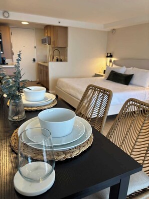A dining table with white plates, bowls, and glasses, a wicker chair, and a bed with white linens in the background. - Newly Renovated, Chic Studio w/ Ocean Views (Honolulu)