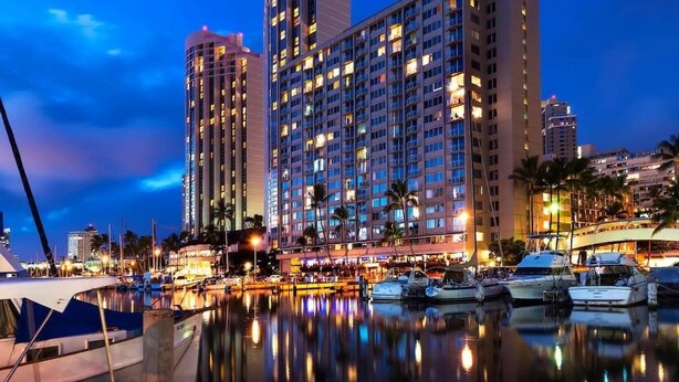 A marina with boats docked, a large hotel building with multiple floors, and a clear sky. - Marina Front, Large Lanai, Near Beach (Honolulu)