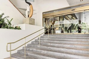 A staircase with a metal handrail, a white wall, and a glass door leading to an outdoor area with plants. - Bright Central Studio at Luana Hotel + Suites (Honolulu)