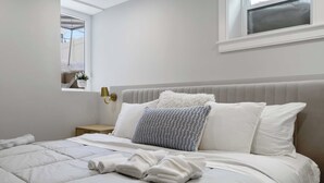 Ein ordentlich bezogenes Bett mit weißer Bettwäsche und einem flauschigen Kopfkissen, ein Nachttisch mit Lampe und ein Fenster mit Blick ins Freie.