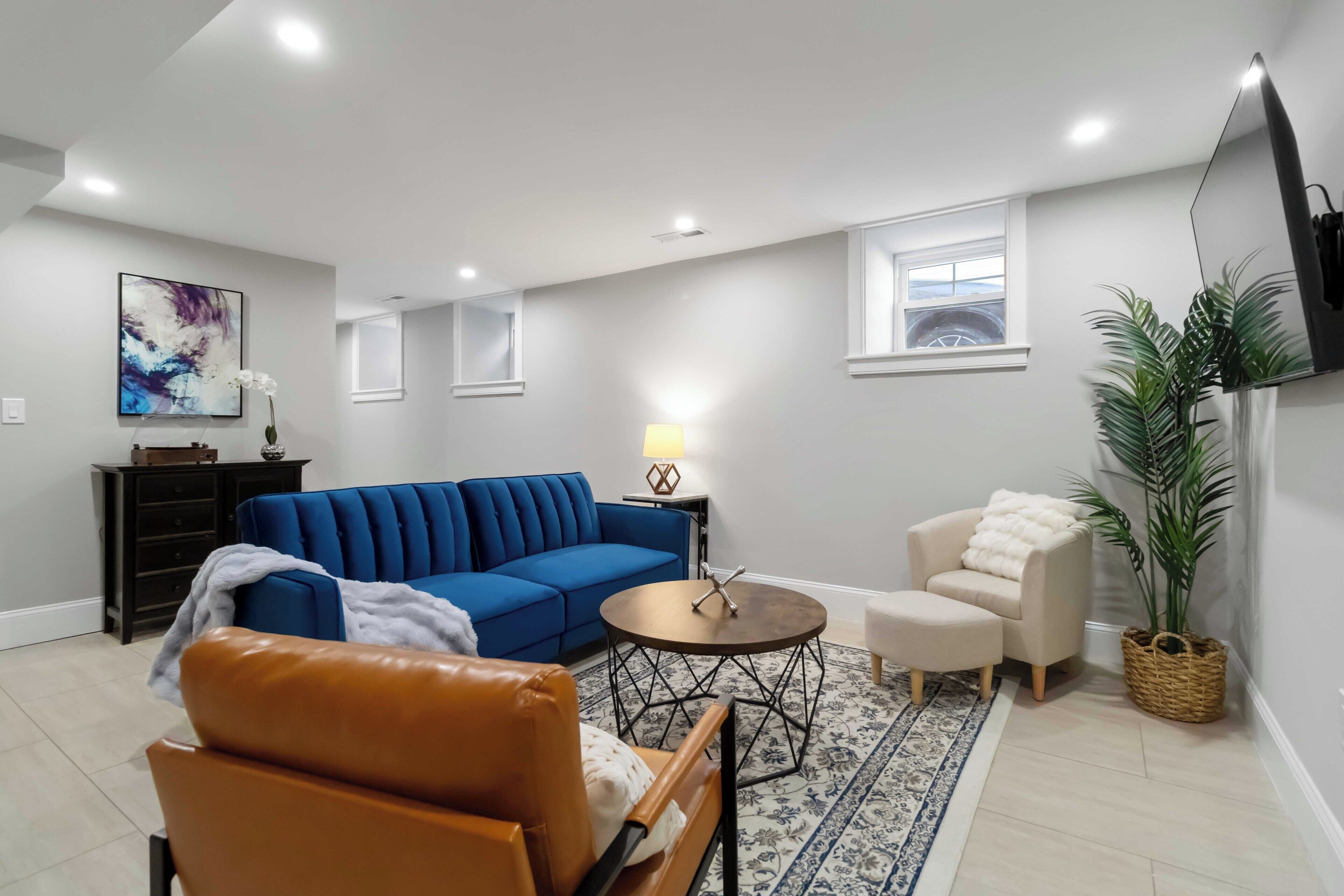 A living room with a blue sofa, a brown leather chair, a small round table, a lamp, a white armchair, a large potted plant, and a TV mounted on the wall.