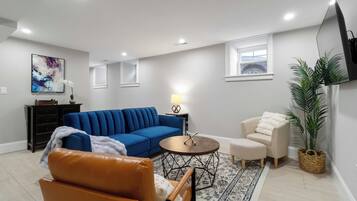 A living room with a blue sofa, a brown leather chair, a small round table, a lamp, a white armchair, a large potted plant, and a TV mounted on the wall.