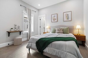 A bedroom with a bed, bedside lamps, a nightstand, a window with curtains, a shelf with decorative items, and a door.