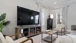Ein Wohnzimmer mit Flachbildfernseher, einem Holztisch, einem beigefarbenen Sofa, einem Sessel und einer großen Pflanze.