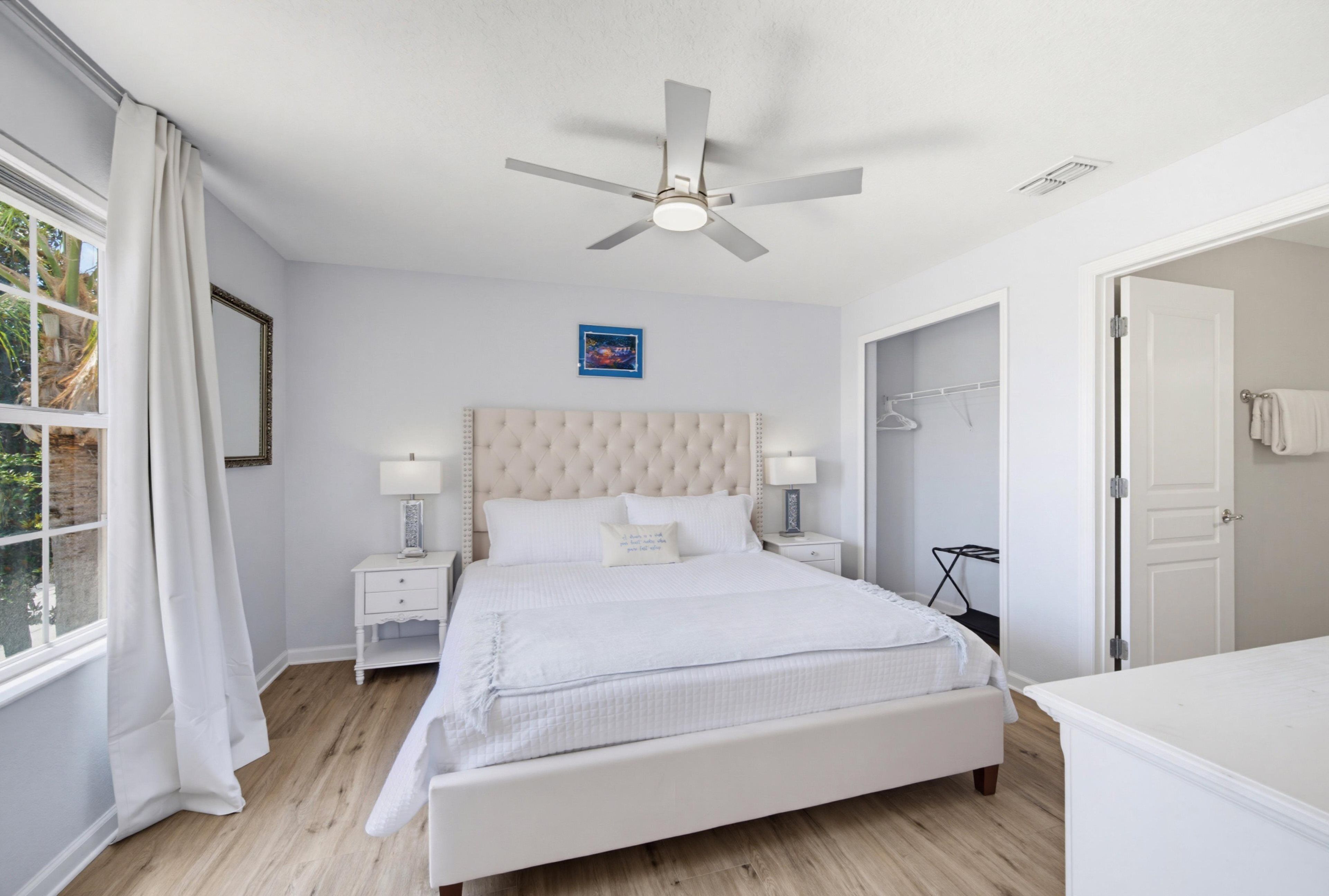 A bedroom with a large bed, a ceiling fan, a window with curtains, a bedside table with lamps, and a door leading to another room.