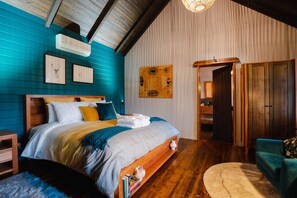 A bedroom with a wooden bed, a blue wall, a wooden floor, a blue armchair, and a door leading to another room. - The perfect spot for family and friends (Bright)