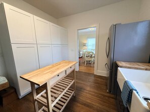 A kitchen with white cabinets, a wooden table, a refrigerator, and a dining area in the background.