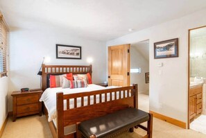 A bedroom with a wooden bed, bedside tables, a bench, a wardrobe, and a bathroom with a mirror and sink. - Telluride Trails Retreat: 3BR/2BA - Hot Tub/Sauna (Telluride)