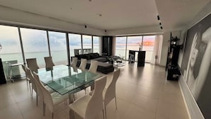 Een moderne woonruimte met een glazen eettafel, witte stoelen, een bank en uitzicht op zee.