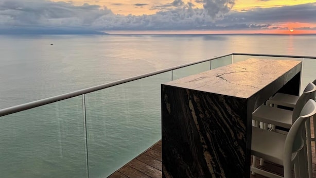 Een balkon met een marmeren tafel en stoelen, met uitzicht op een kalme zee bij zonsondergang.