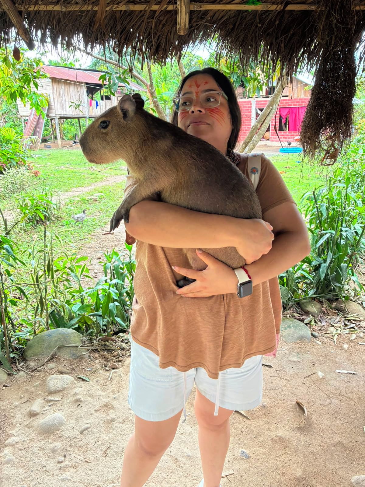 A person holding a capybara in a rural setting with a thatched roof structure and greenery.