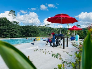 A pool area with a sandy surface, red umbrellas, and people swimming and relaxing. - Animal Glamping (Pomona)