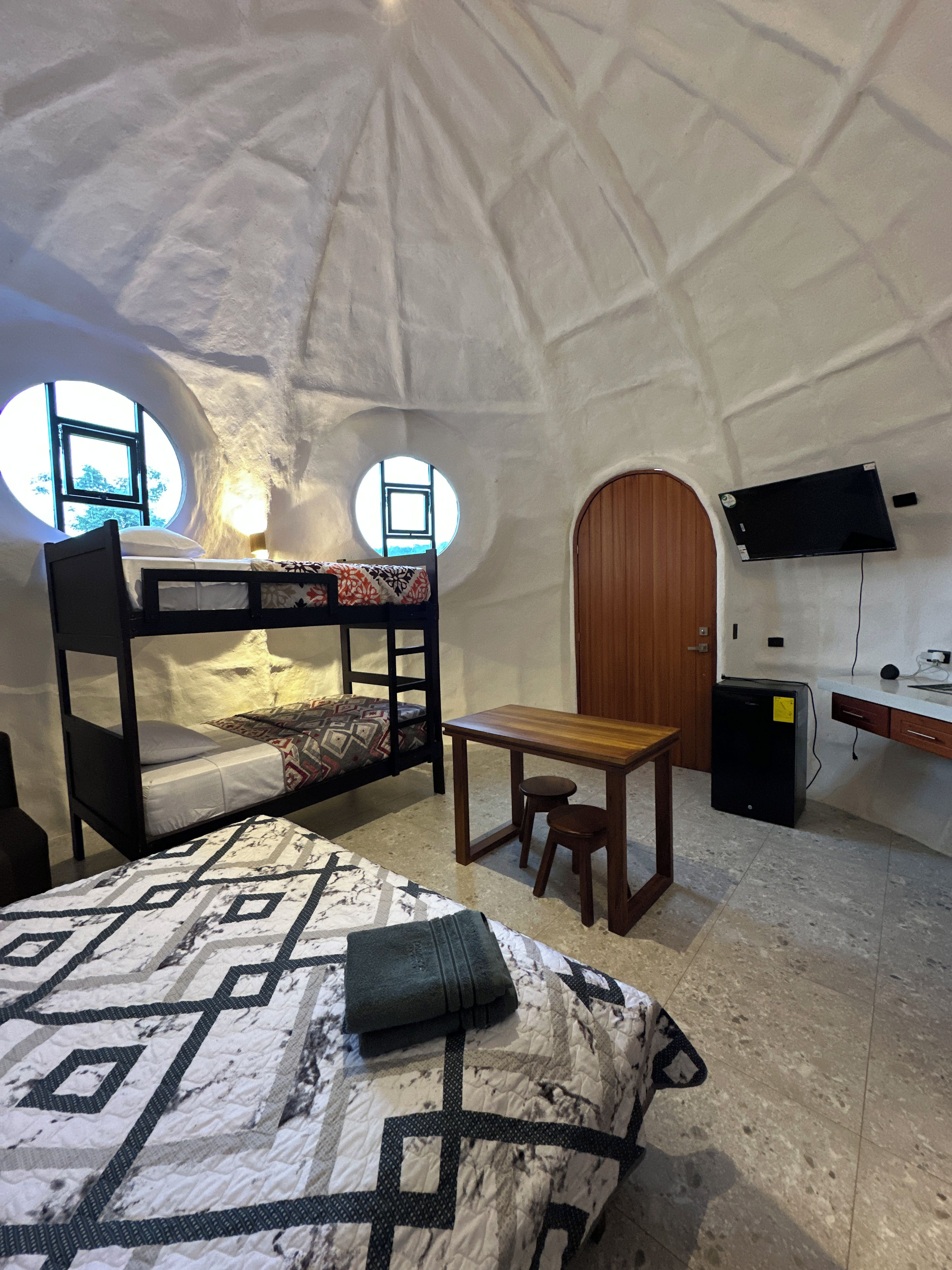 Interior acogedor de una estructura en forma de cúpula, con una cama, un escritorio, una silla, un televisor montado en la pared y un refrigerador pequeño.