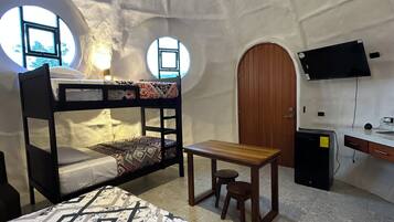 Interior acogedor de una estructura en forma de cúpula, con una cama, un escritorio, una silla, un televisor montado en la pared y un refrigerador pequeño.