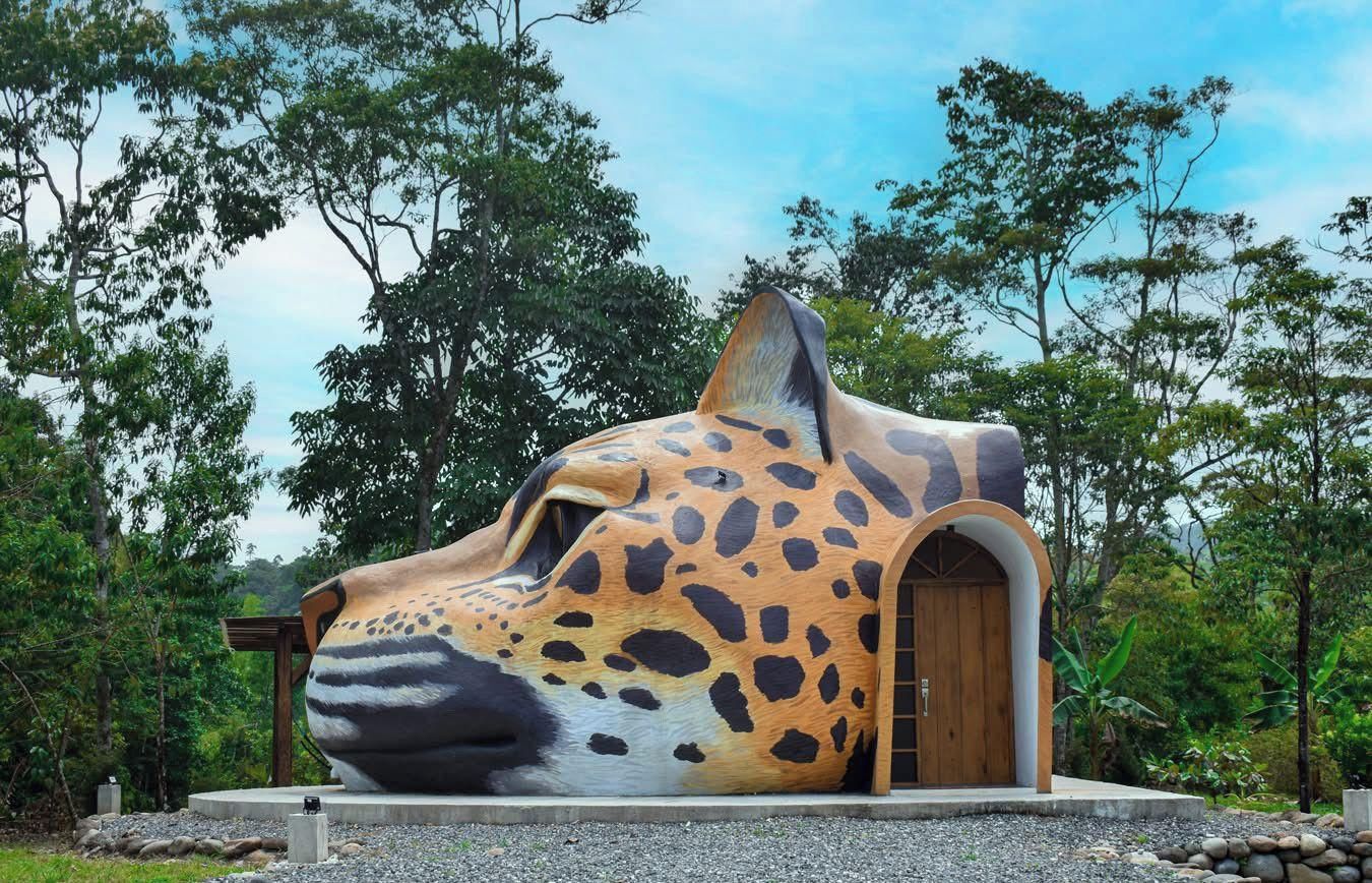 Eine große, skulpturale Struktur, die einen Jaguar-Kopf mit einem Bogenportal darstellt, eingebettet in eine natürliche Umgebung mit Bäumen und blauem Himmel.