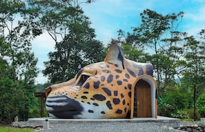 A large, sculptural structure resembling a jaguar's head with an arched entrance, set in a natural environment with trees and a clear sky. - Animal Glamping (Pomona)