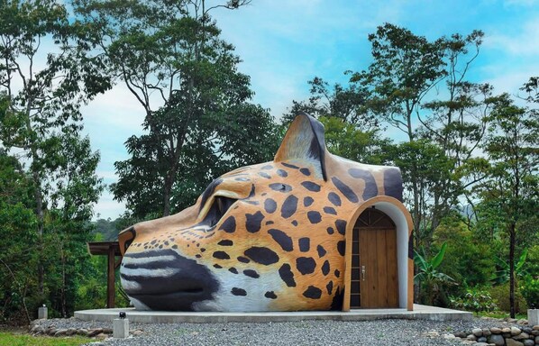 A large, sculptural structure resembling a jaguar's head with an arched entrance, set in a natural environment with trees and a clear sky. - Animal Glamping (Pomona)
