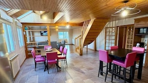 A spacious room with a wooden staircase, a dining area with pink chairs, and a kitchen area. - Gîte du Roc 8/10 persons (SAINT-SAUVEUR-DE-PEYRE)