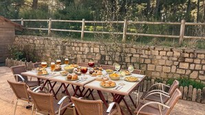 An outdoor dining area with a stone wall, wicker chairs, and a table set with various dishes and drinks. - Gîte du Roc 8/10 persons (SAINT-SAUVEUR-DE-PEYRE)