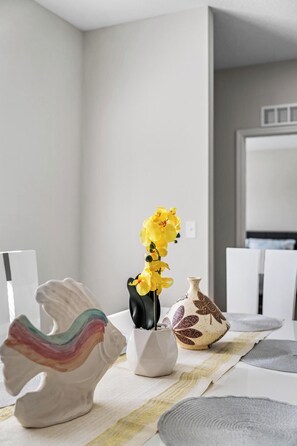 Ein Esstisch mit weißer Tischdecke, eine farbenfrohe Keramikpferd-Skulptur, eine Vase mit gelben Blumen und ein verziertes Keramikgefäß.