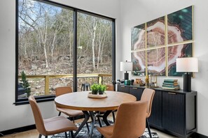 A dining area with a round table, chairs, a view of trees through large windows, and a cabinet with decorative items. - Glass Cabin Over Creek | Dome+Hot Tub+Grotto Pool (Omaha)