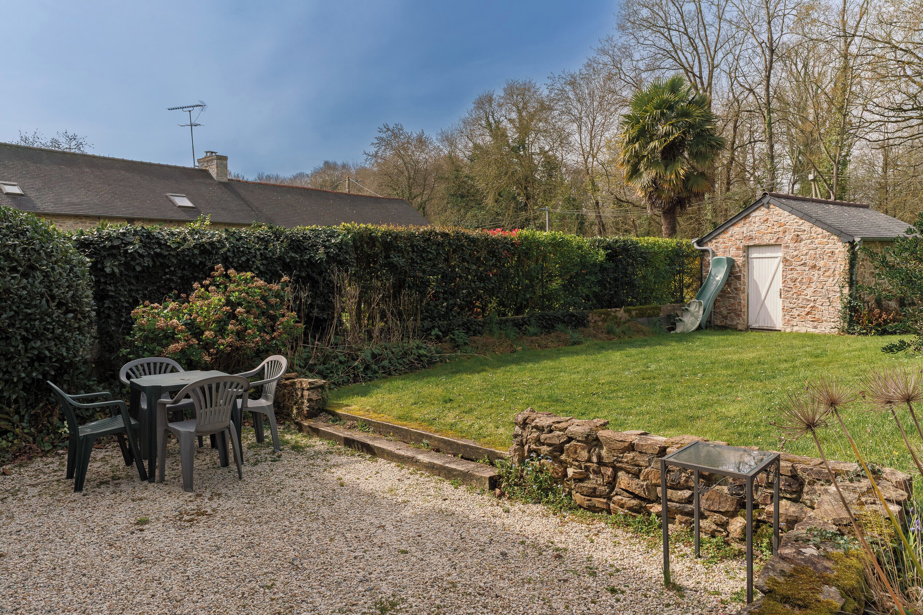 A gravel patio with a table and chairs, a stone wall, a small stone building with a door and a slide, and a hedge.