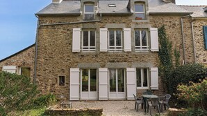 A stone building with multiple windows and shutters, a small outdoor seating area with chairs and a table, and a gravel pathway leading to the entrance.