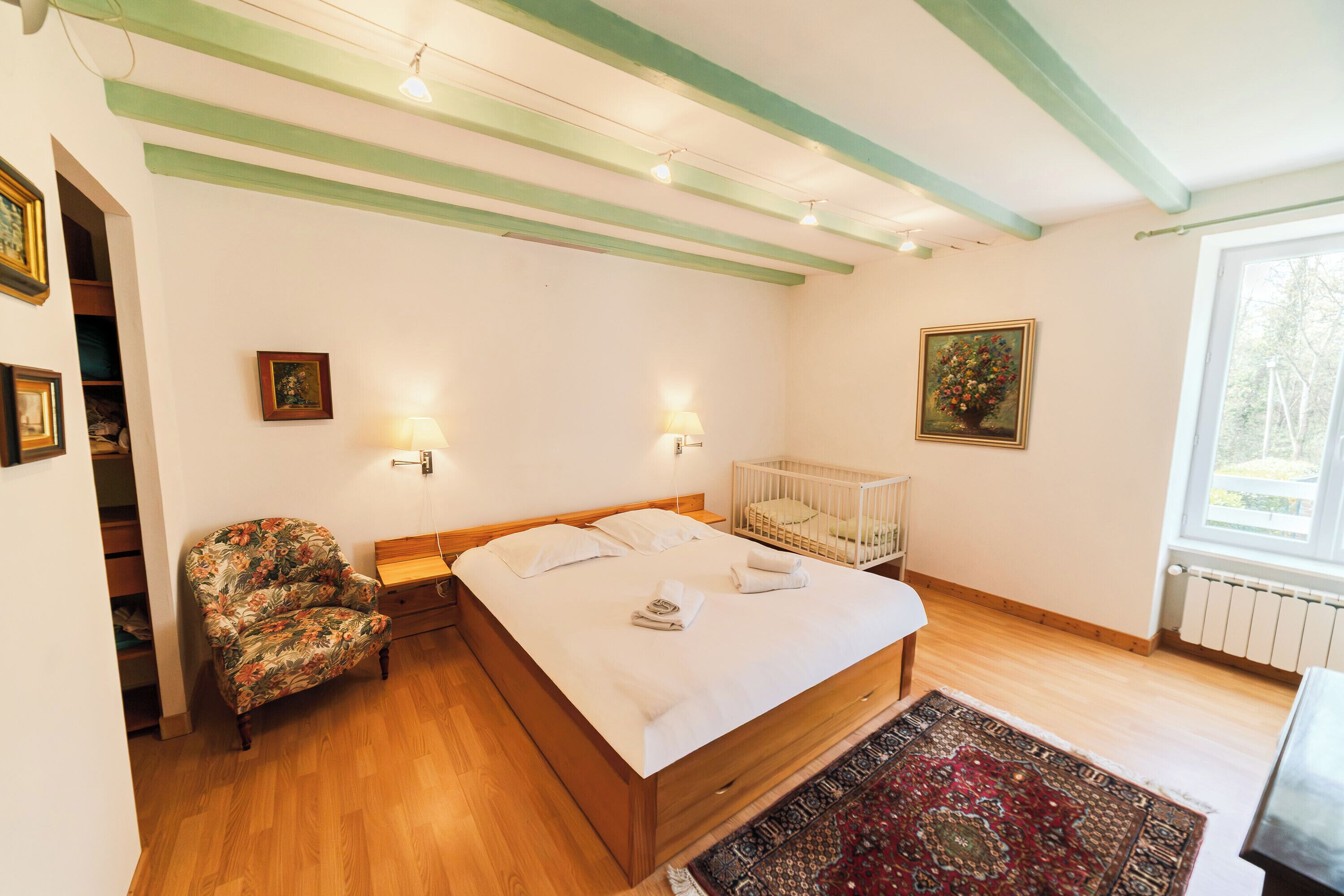 Une chambre à coucher avec un lit en bois, un fauteuil à motifs floraux, un berceau et une vue sur l’extérieur par une fenêtre.