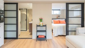 Ett modernt hotellrum med dusch, en säng med orange kuddar, en soffa och ett litet bord med en vas.