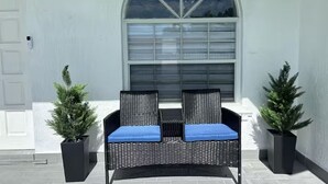 A black wicker bench with blue cushions in front of a white door, flanked by two potted evergreen plants. - Miami Retreat with Backyard Pool (Miami)