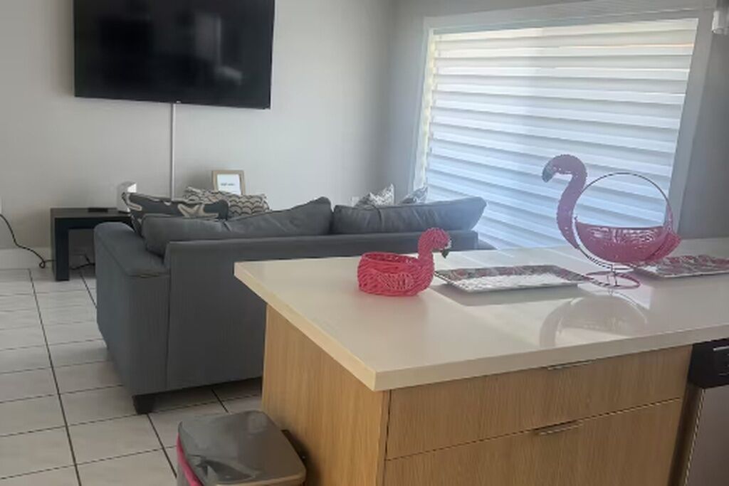 Una sala de estar con sofá, televisión montada en la pared, ventana con persianas y una pequeña cocina con mostrador.