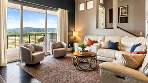 A spacious living room with a large sliding glass door leading to a balcony, a white leather sofa, two armchairs, a coffee table, and a lamp.