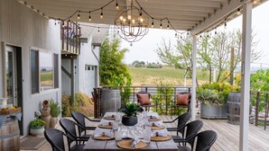 An outdoor patio with a wooden table set for dining, surrounded by wicker chairs, and a view of a vineyard in the background.