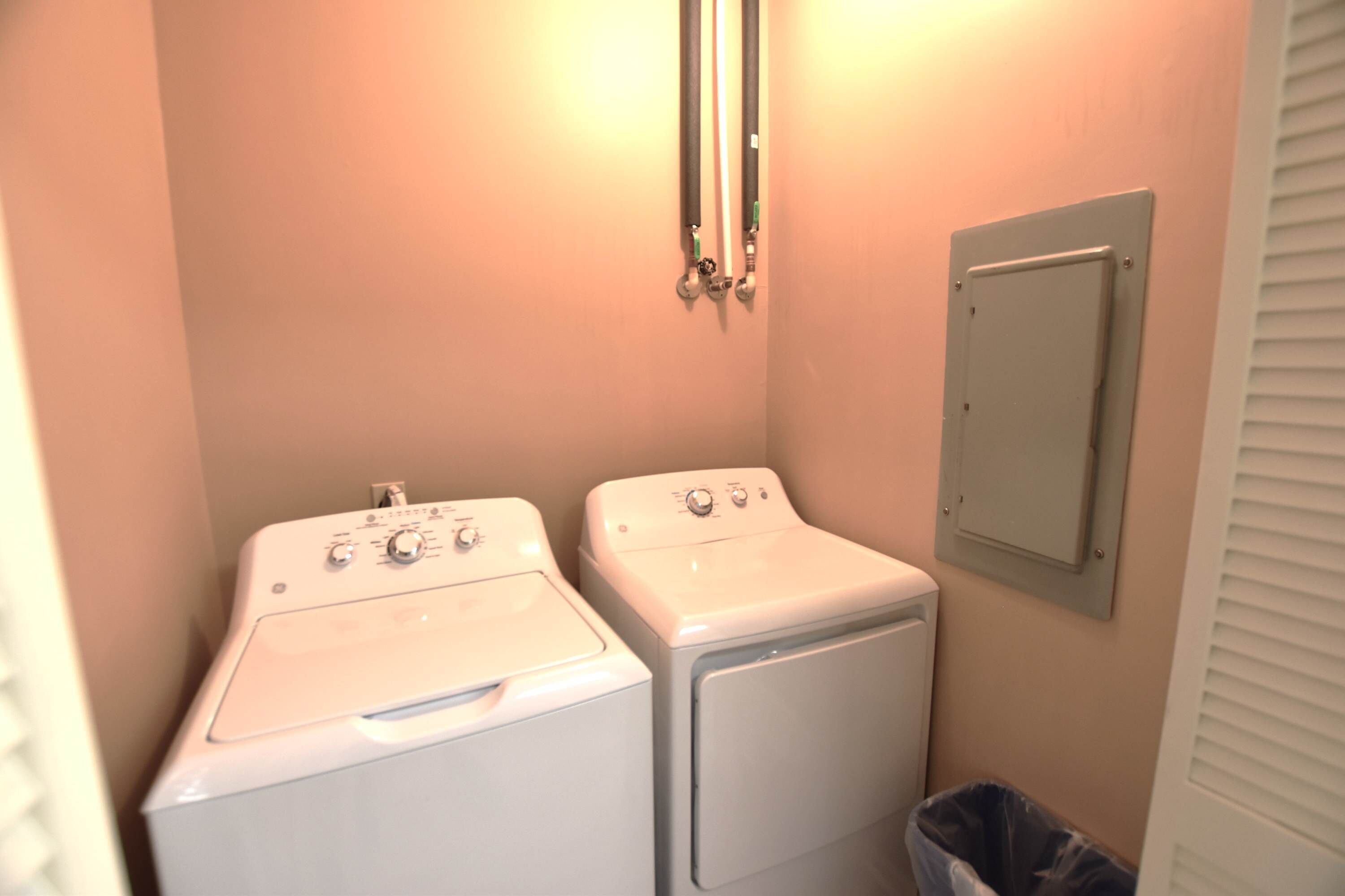 A laundry room with two washing machines, a wall-mounted heater, and a trash bin.