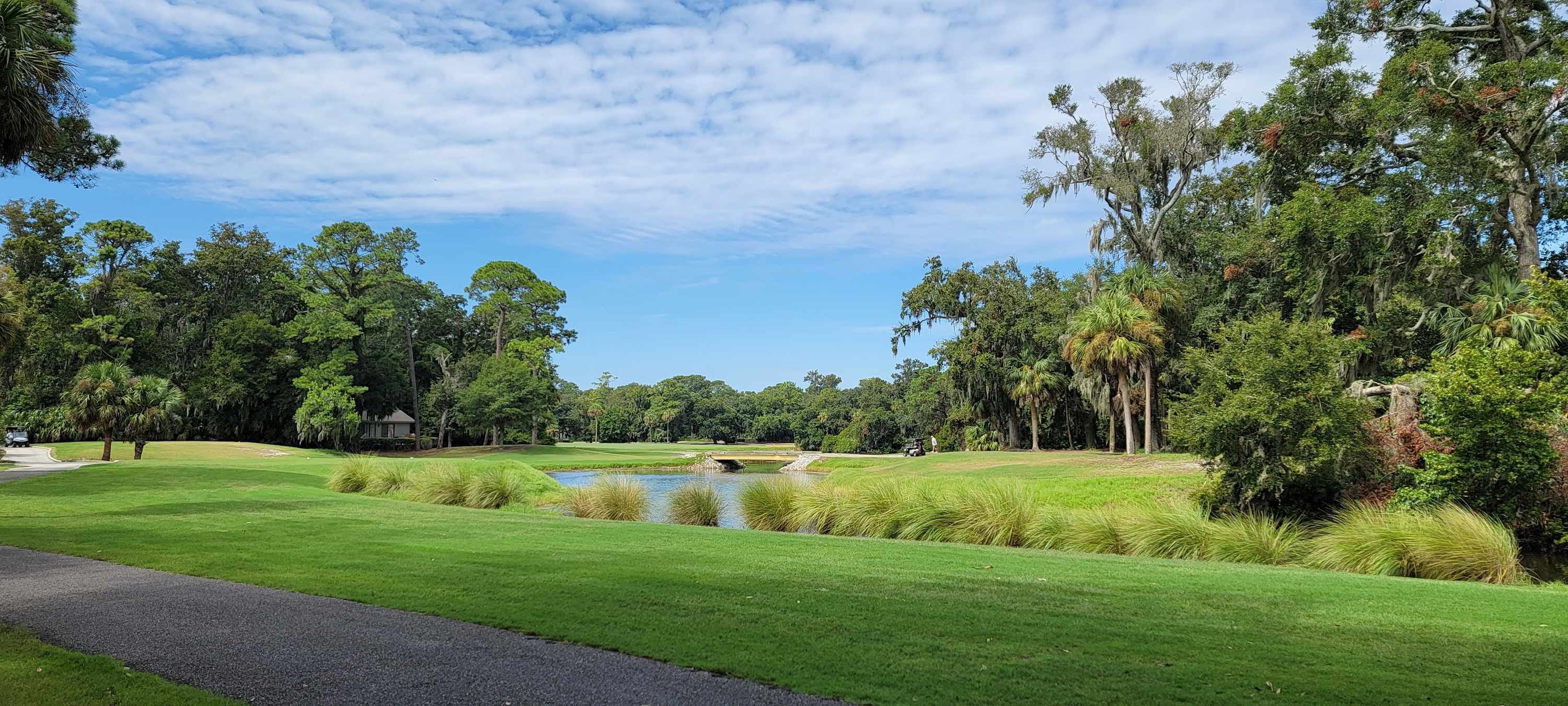 Un campo de golf con un estanque, abundante vegetación y un cielo despejado.