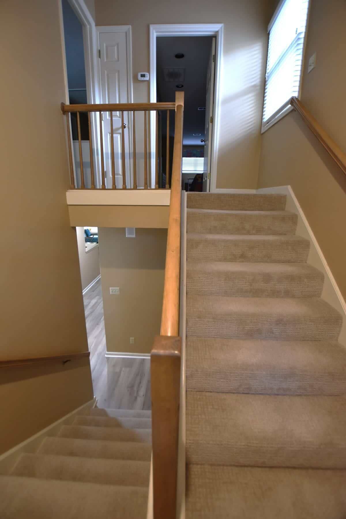 Eine Treppe mit hölzernem Geländer, Teppichstufen und Blick auf einen Raum im zweiten Stock mit Tür und Fenster.