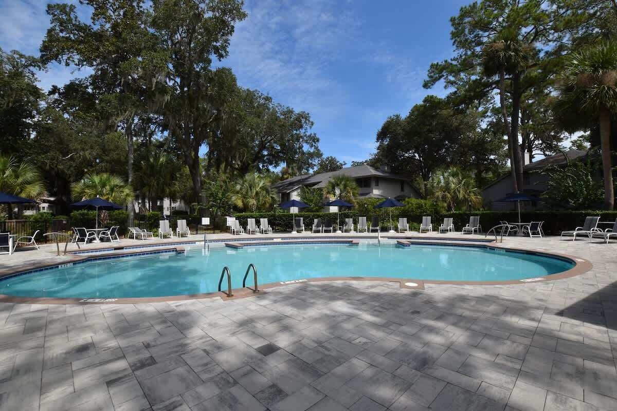 A swimming pool with lounge chairs and umbrellas, surrounded by trees and a building in the background.