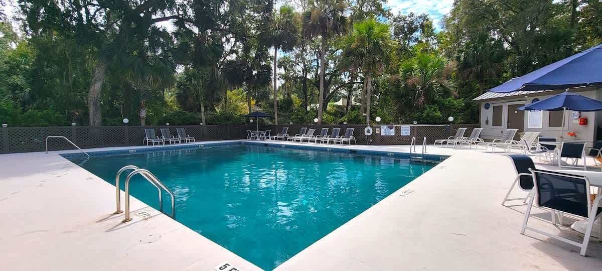 Et rektangulært swimmingpool med en metalstige, omgivet af liggestole og parasoller, med træer i baggrunden.