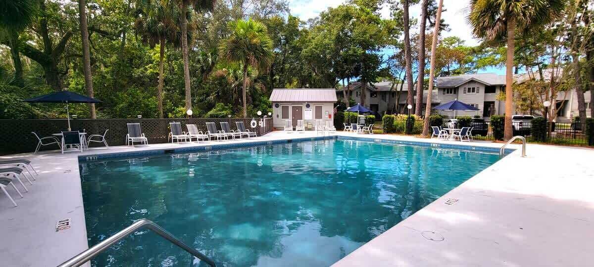 A swimming pool with lounge chairs and umbrellas, surrounded by trees and a building.