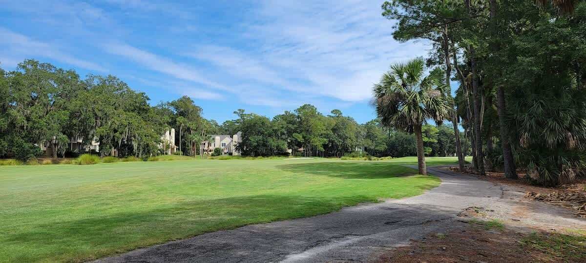 A golf course with a clear path, surrounded by trees and buildings in the background.