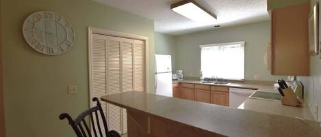 A kitchen with a bar counter, a clock, and a window with blinds.