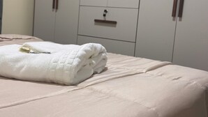 Una cama con un edredón blanco y una toalla blanca doblada. Se observa un armario de madera con dos puertas y un cajón pequeño al fondo.
