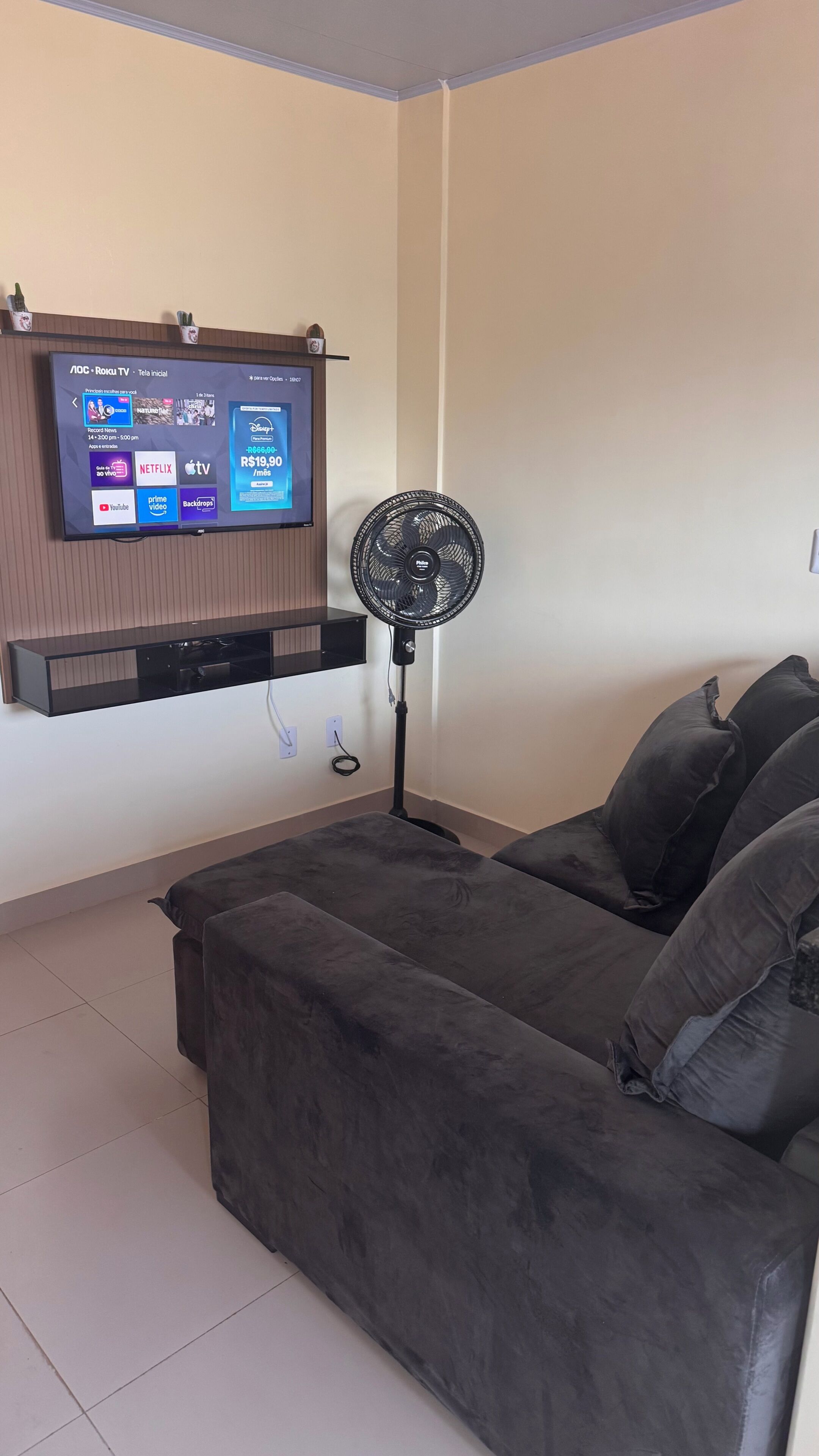 A living room with a flat-screen TV mounted on the wall, a standing fan, a grey sofa, and a small shelf with decorative items.
