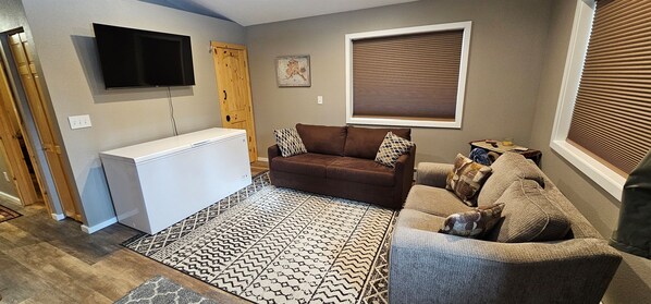 Un salon avec un canapé marron, un canapé gris, un tapis à motifs, un téléviseur fixé au mur et une fenêtre avec des stores.