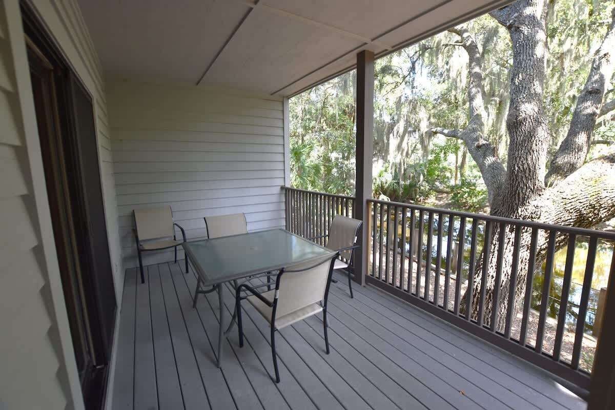 A covered porch with a table and chairs, surrounded by a railing, overlooking a wooded area.